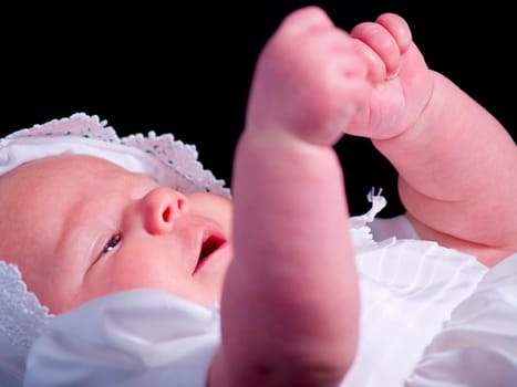 A portrait of a baby girl in a christening dress on a dark background. This photo has a shallow depth of field and the focus is on the left hand.