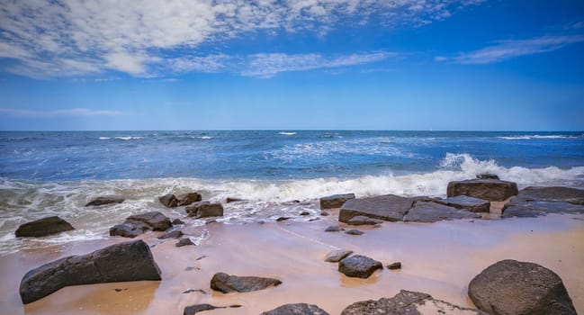 Rocky beach at Alexandra Headland, Maroochydore, Sunshine Coast, Queensland. The tide is coming in, sand is wet and surf waves breaking over the rocks. Blue sky with clouds and blue ocean.