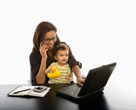 African American mid adult businesswoman working on laptop and cell phone with toddler son on lap.