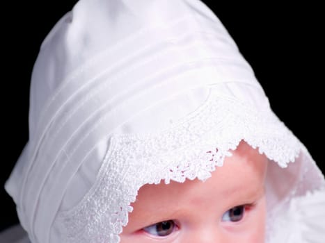 A portrait of a baby girl in a christening dress on a dark background. This photo has a shallow depth of field and the focus is on the edge of her hood next to her eye..