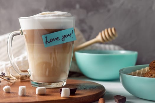 Coffee latte cup with Love you sticker on wooden tray with turquoise bowls with cookies and marshmallows on gray wall background