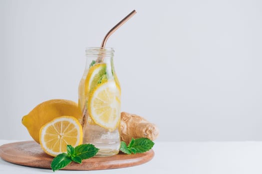 Glass bottle with lemonade with metal straw and ingredients for cooking on wooden tray. Ginger, lemon, mint on white background. Copy space
