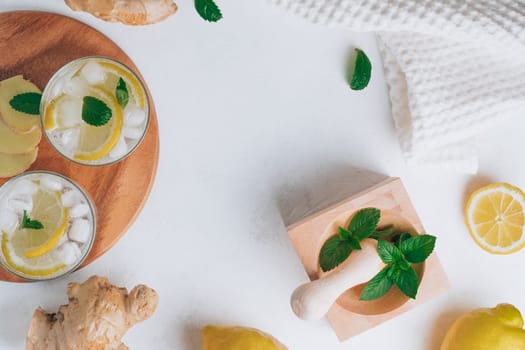 Two glasses with lemonade and ingredients for cooking. Ginger, lemon, mint, ice on white surface. Wooden tray, mortar and pestle. Flat lay. Copy space
