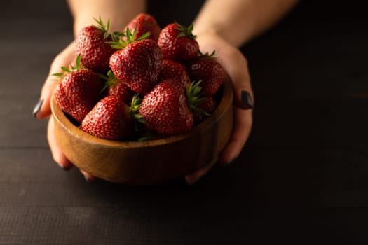 Hands holding a wooden bowl of fresh red ripe strawberries on a black wooden surface. Low key. Copy space.
