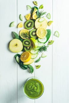 Glass of green smoothie with vegetables, fruits and herbs it is made of, on wooden white surface. Flat lay, vertical