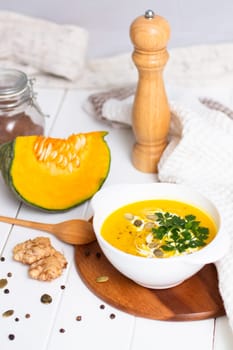 A plate of yellow pumpkin cream soup with cream, herbs and seeds on a white table near ginger, raw pumpkin and a wooden spoon. Vertical