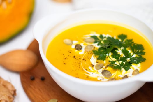 A plate of yellow pumpkin cream soup with cream, herbs and seeds on a white table near ginger, raw pumpkin and a wooden spoon.