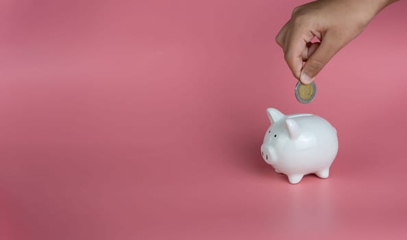 A human hand is dropping a coin into a white piggy bank. on a pink background. Savings and investment concepts.