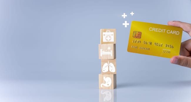 Hand holding credit card and wooden cube blocks stacked with icons. Concept of medical expenses, health and medicine.
