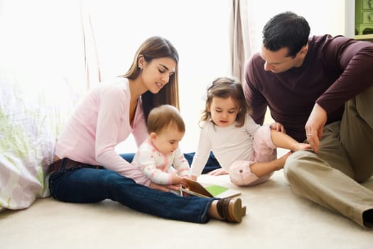 Caucasian girl children with mother and father sitting on bedroom floor looking at book.