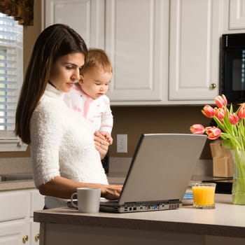 Mother holding baby  and typing on laptop computer in kitchen.