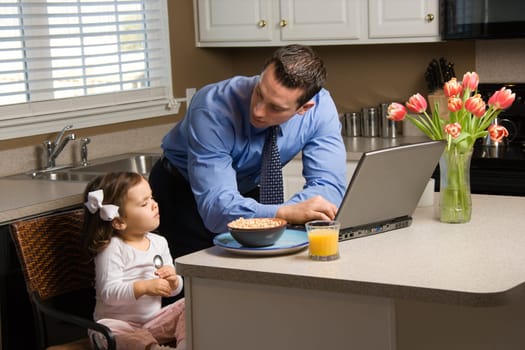 Caucasian father in suit using laptop computer with daughter eating breakfast in kitchen.