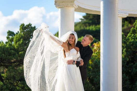 Happy newlyweds hugging on a walk, the girl raised her veil with her hand and smiles