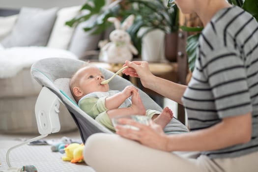 Mother spoon feeding her baby boy child in baby chair with fruit puree. Baby solid food introduction concept