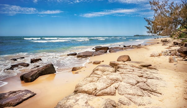 Ebb tide coming in on a rocky seashore. Surf waves break over shallow reef. Cloudy blue sky on a spring day on Sunshine Coast, Queensland.