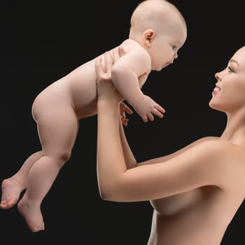 A mother joyfully lifts her baby while both share a playful and loving expression, showcasing a bond in a serene setting with a dark background.