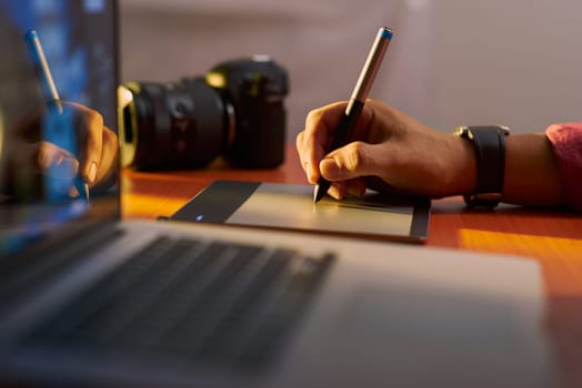 Photographer drawing and retouching image on laptop computer, using a digital tablet and stylus pen. Closeup of man's hand with dslr camera in background. Copy space in foreground
