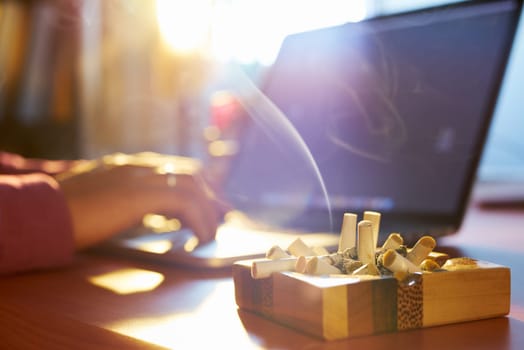 Close up of ashtray full of cigarette, with man in background working on laptop computer and smoking indoors on early morning. Concept of addiction and abuse of nicotine.