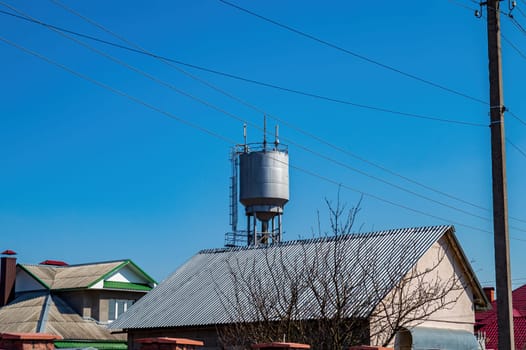 A water tower over the roofs of rural houses against the blue sky. Water tower. Roofs of houses. Blue sky. Water supply in the village. Metal industrial construction. Background image.