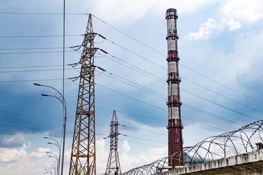High voltage tower with wires and industrial chimney. High-voltage transmission line of electrical energy. Chimney of the enterprise. Industrial landscape. Cloudy sky horizon. Background.