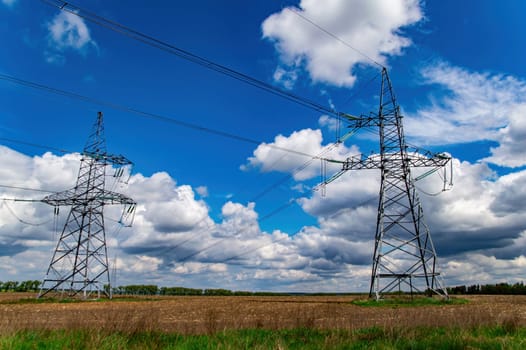 High-voltage power transmission tower with wires. High voltage line. Energy industry. Energy transfer. Blue sky. White clouds. Agricultural field. Cloudy horizon. Business. Electrician work.