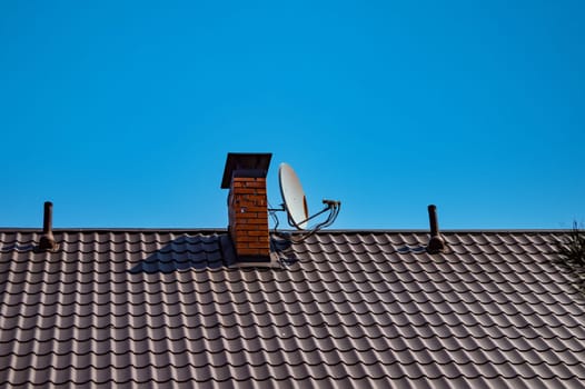 Satellite television antenna attached to the chimney of a rural house. Satellite TV antenna. Chimney at home. Tile roof. Blue sky. Modern technologies. Broadcasting. Electronic technologies.