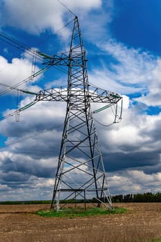High-voltage power transmission tower with wires. High voltage line. Energy industry. Energy transfer. Blue sky. White clouds. Agricultural field. Cloudy horizon. Business. Electrician work.
