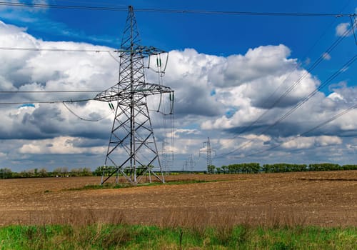 High-voltage power transmission tower with wires. High voltage line. Energy industry. Energy transfer. Blue sky. White clouds. Agricultural field. Cloudy horizon. Business. Electrician work.