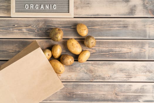 Bunch of unpeeled yellow new potatoes on faded wooden table, top view. Fresh farm vegetables take out of eco paper bag. Organic food or healthy diet concept. Planking. Culinary and cooking.