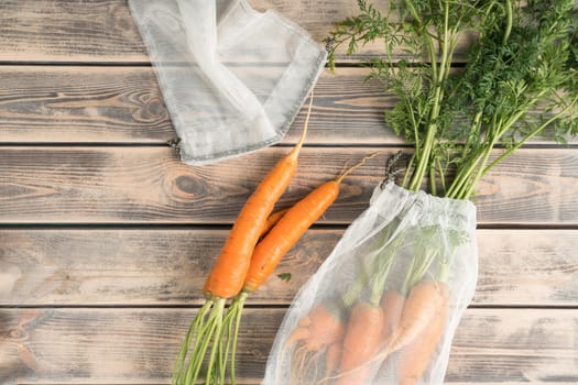 Bunch of fresh ripe carrots with long green haulm on faded wooden table, top view. Unpacking vegetables from transparent fabric bags. Organic food or healthy diet concept. Objects layout. Planking.