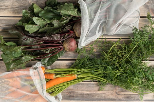 Bunches of fresh beets and carrots with long green haulm on wooden table, top view. Unpacking farm vegetables from eco fabric bags. Organic food or healthy diet concept. Objects layout. Planking.