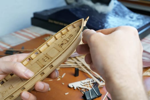 Hands of man cutting out details for ship model with clerical knife from plywood. Process of building toy ship, hobby and handicraft. Table with various materials, parts and devices for work