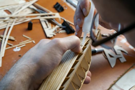 Hands of man gluing plywood details for ship model with glue, holding with fingers. Process of building toy ship, hobby, handicraft. Table with various materials, parts and devices for work