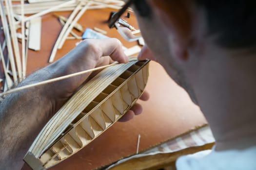 Hands of man gluing plywood details for ship model with glue, holding with fingers. Process of building toy ship, hobby, handicraft. Table with various materials, parts and devices for work