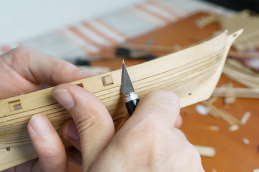 Hands of man cutting out details for ship model with clerical knife from plywood. Process of building toy ship, hobby and handicraft. Table with various materials, parts and devices for work