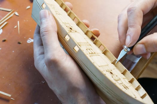 Hands of man cutting out details for ship model with clerical knife from plywood. Process of building toy ship, hobby and handicraft. Table with various materials, parts and devices for work