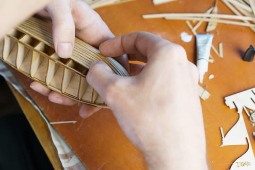 Hands of man gluing plywood details for ship model with glue, holding with fingers. Process of building toy ship, hobby, handicraft. Table with various materials, parts and devices for work