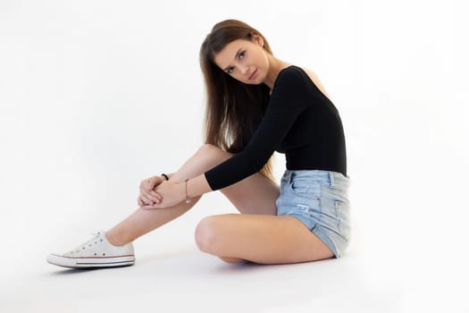 Studio shot of young long haired and long legged brunette woman sitting on floor and looking at camera. Horizontally.