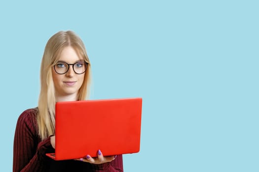 Image of cheerful young woman standing isolated over blue background using red laptop computer. The girl is looking at the camera.