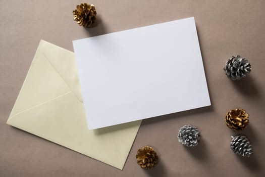 Christmas winter festive ornament. Blank paper card and envelope with silver and golden pine cones on brown background. Minimal decoration. Invitation Christmas holiday party. Flat lay, top view, copy space.