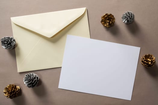 Christmas winter festive ornament. Blank paper card and envelope with silver and golden pine cones on brown background. Minimal decoration. Invitation Christmas holiday party. Flat lay, top view, copy space.