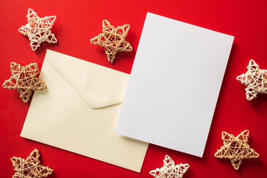 Christmas winter festive ornament. Blank paper card and envelope with wooden decoration on red background. Minimal decoration. Invitation Christmas holiday party. Flat lay, top view, copy space.