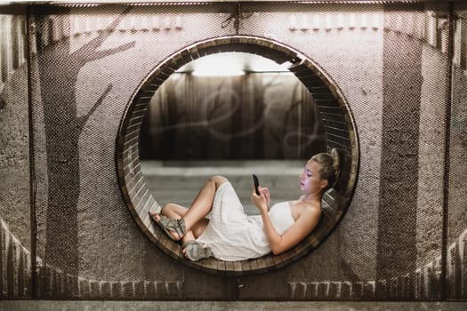 Lady wearing white summer dress playing carelessly with her android smart phone browsing trough social networks relaxing on contemporary circular bench in urban pedestrian underpass.