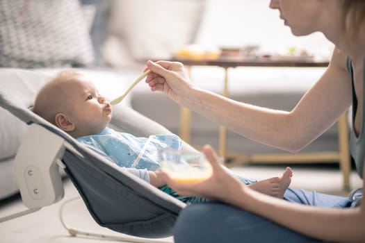 Mother spoon feeding her baby boy child in baby chair with fruit puree. Baby solid food introduction concept