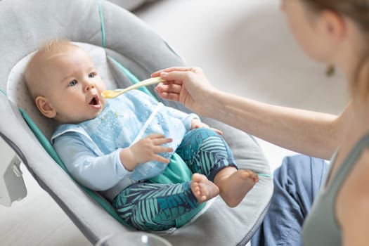 Mother spoon feeding her baby boy child in baby chair with fruit puree. Baby solid food introduction concept