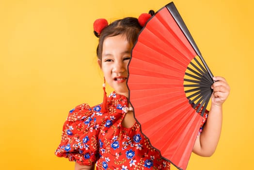 Chinese New Year. Happy Asian Chinese little girl smile wearing red cheongsam holding fan, Portrait children in traditional dress, studio short isolated on yellow background