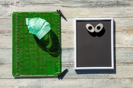 Computer technology background. A green digital electronic circuit board with an old punched tape (memory) on it and a black board with a pair of elliptical gears on a table. Natural light shadows.