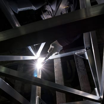 A skilled worker uses a welding torch to join metal beams in a dimly lit workshop, highlighting the intensity of the task.