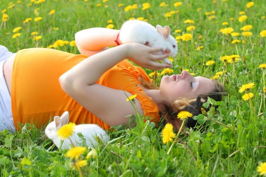 Young pregnant woman lays down on grass and palys with cheerful rabbits