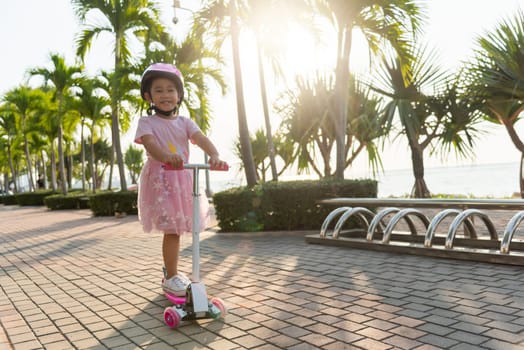 Child riding scooter. Happy Asian little kid girl wear safe helmet playing pink kick board on road in park outdoors on summer day, Active children games outside, Kids sport healthy lifestyle concept
